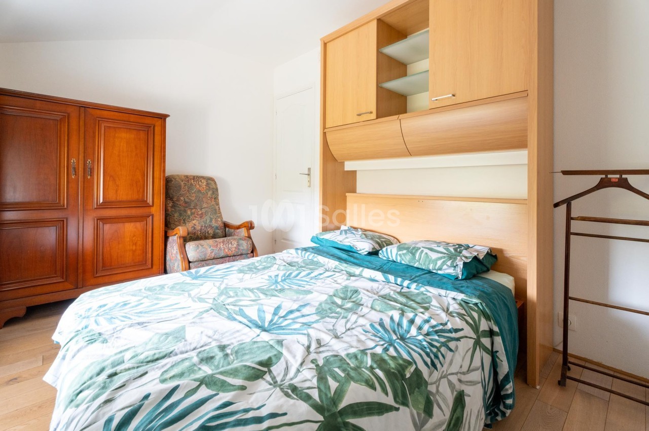Chambre lumineuse avec lit double, housse de couette à motifs végétaux, armoire en bois et fauteuil rembourré.