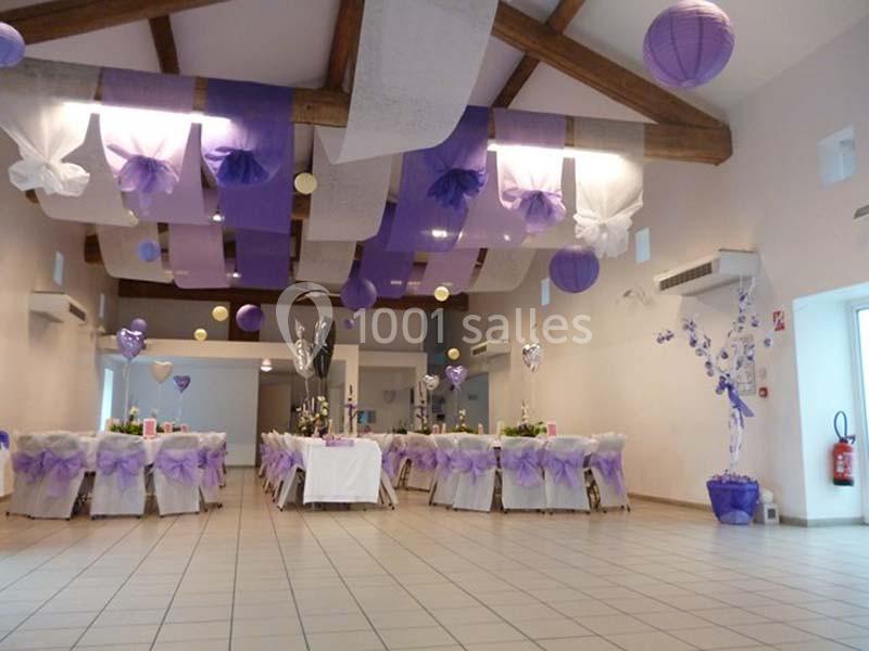 Salle décorée pour un événement avec des tables dressées, des chaises ornées de nœuds violets et des suspensions colorées au…