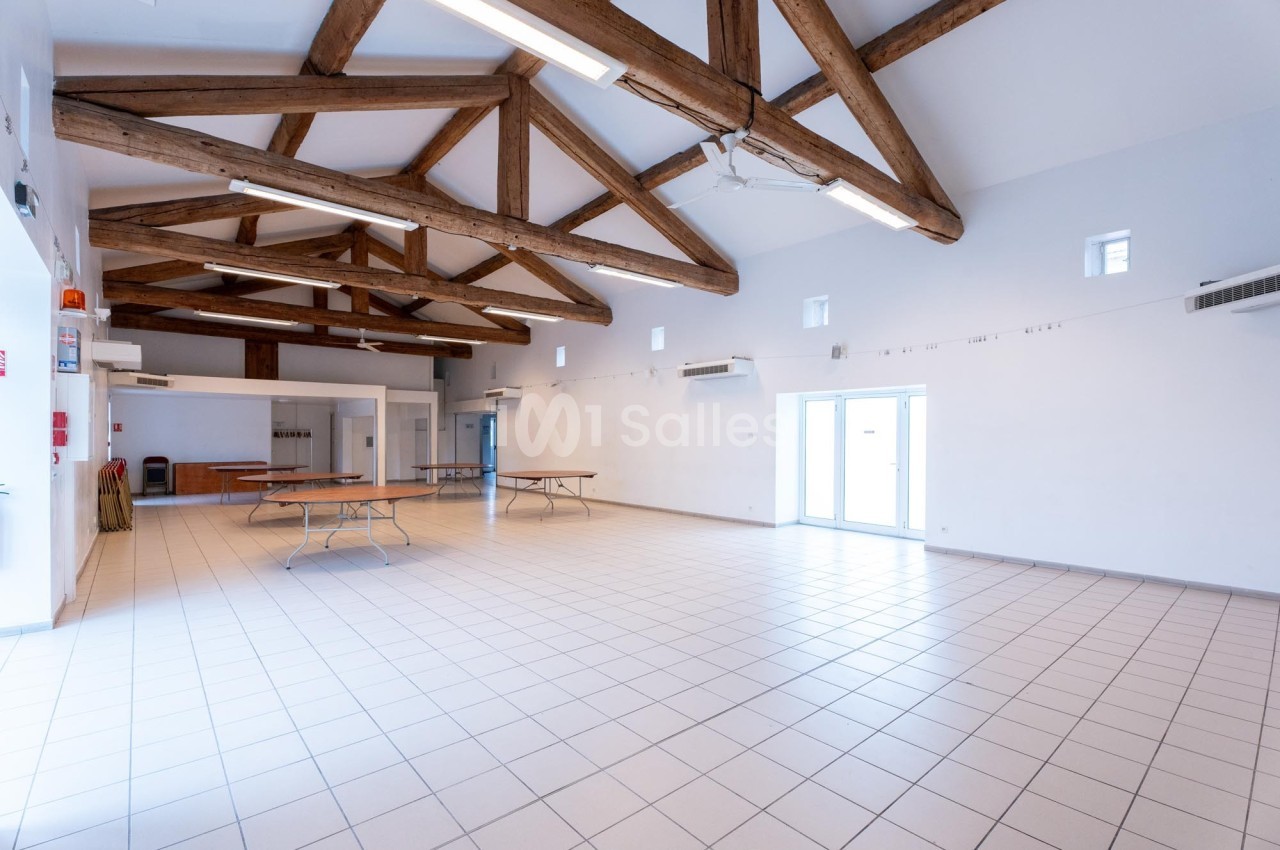 Salle spacieuse avec plafond en poutres apparentes, carrelage clair et tables pliantes contre les murs.
