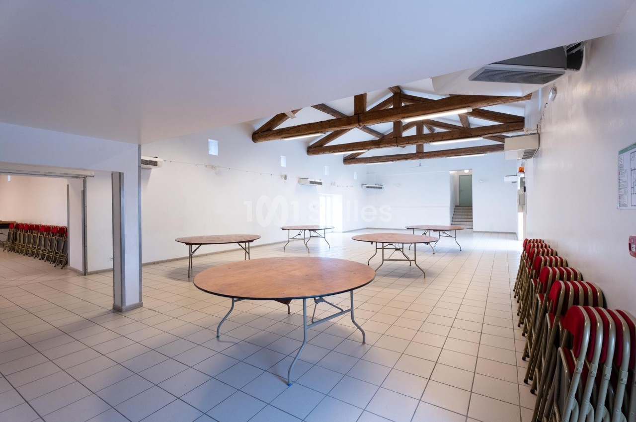 Salle vide avec sol carrelé, tables rondes pliantes espacées et chaises empilées contre les murs, plafond avec poutres…