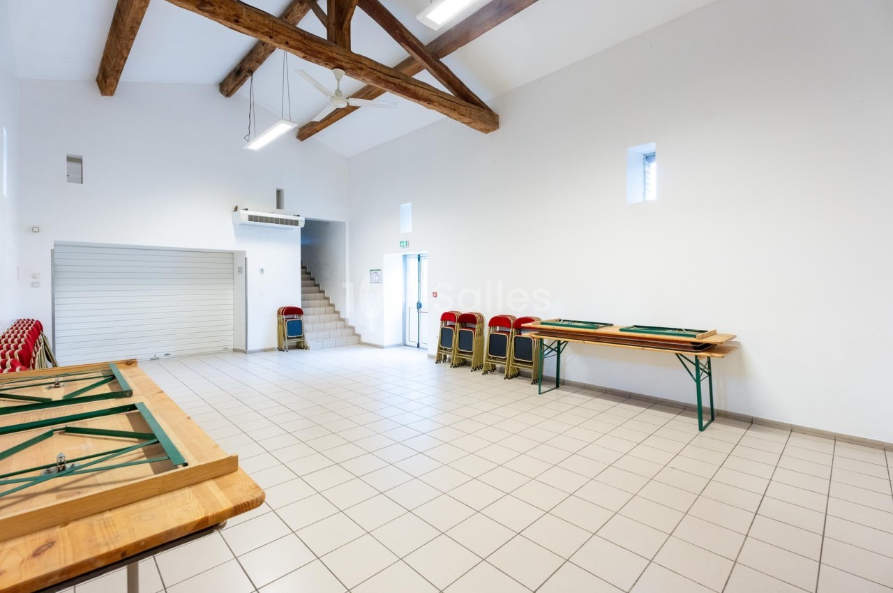 Salle lumineuse avec poutres apparentes, tables pliées, chaises empilées et carrelage clair.