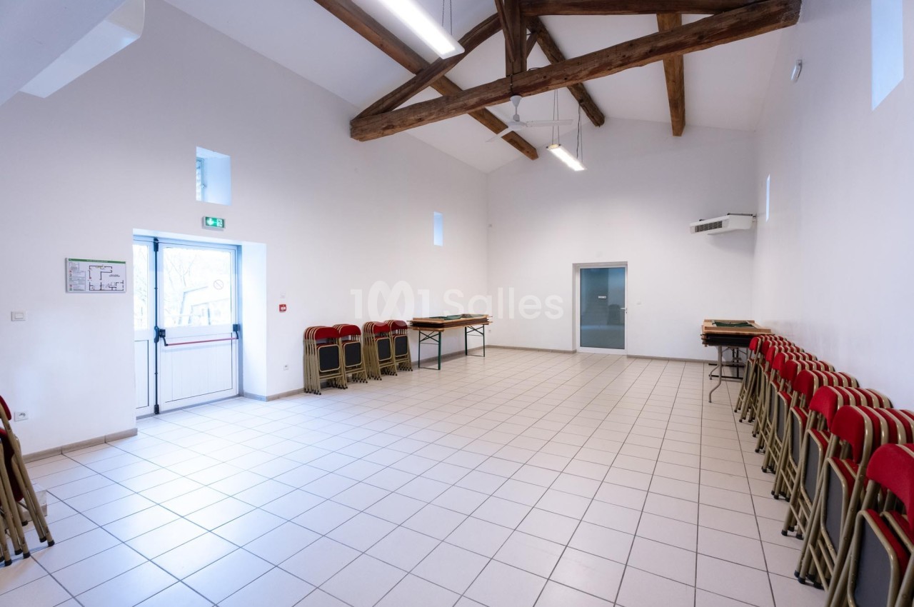 Salle vide avec carrelage clair, poutres apparentes, chaises empilées contre les murs et lumière naturelle.