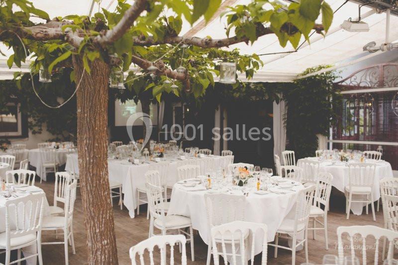 Salle de réception lumineuse avec des tables rondes dressées, chaises blanches et arbres intégrés à la décoration.