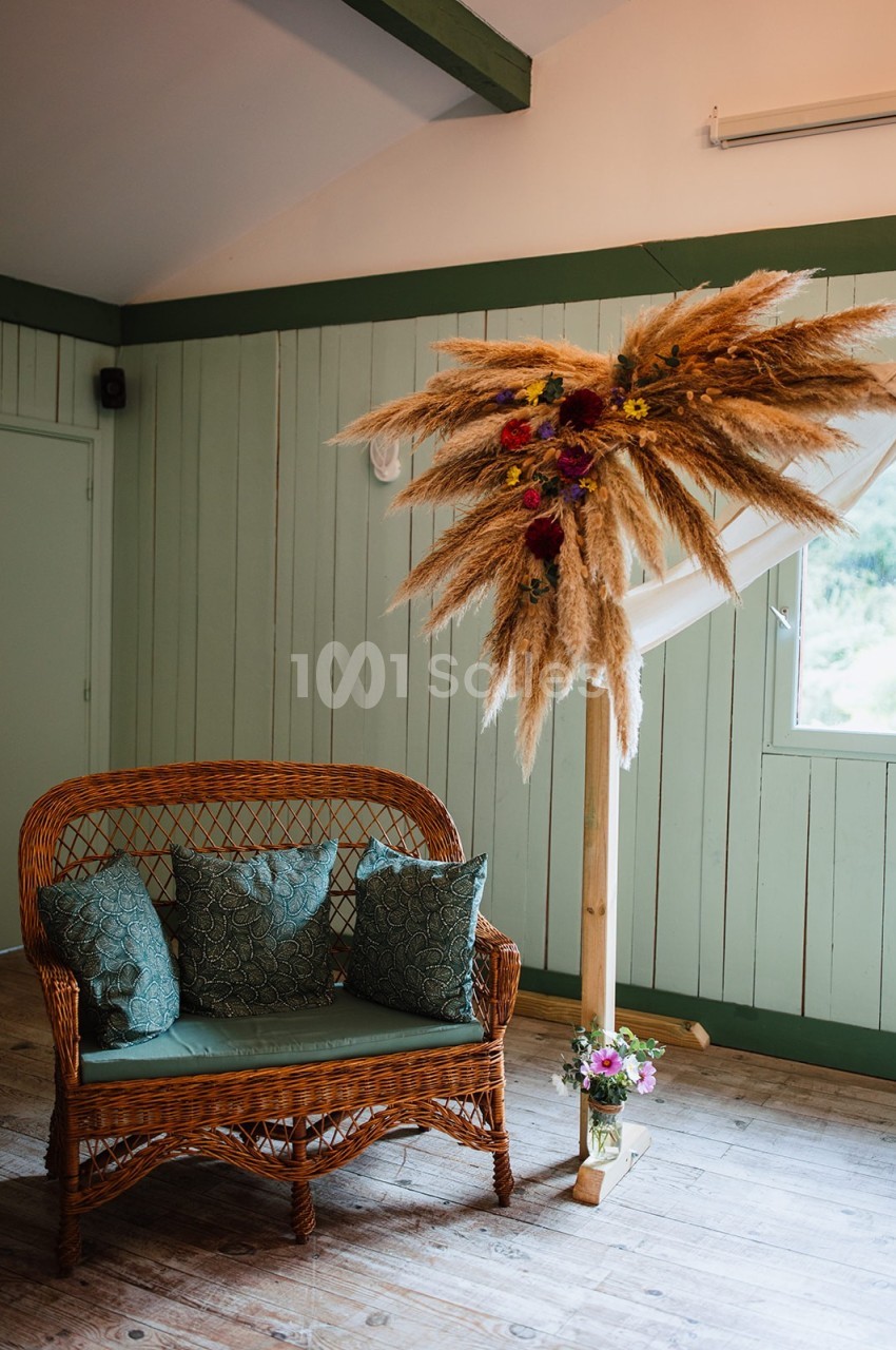 Banc en rotin avec coussins verts, décoré d'une composition florale et de pampas dans une pièce aux murs verts.