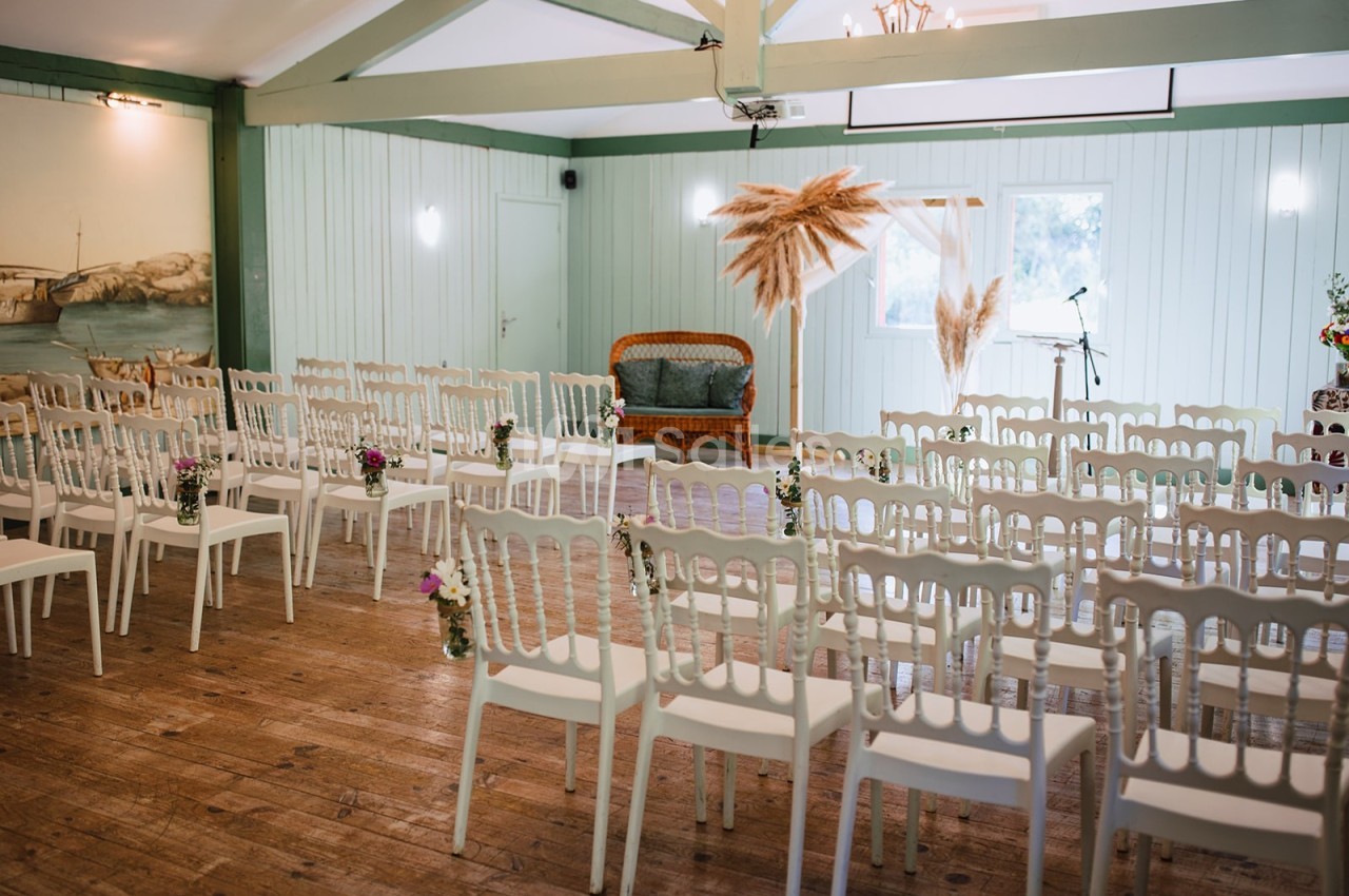 Salle décorée pour une cérémonie avec des chaises blanches alignées, un arche orné de pampas et un canapé en rotin.