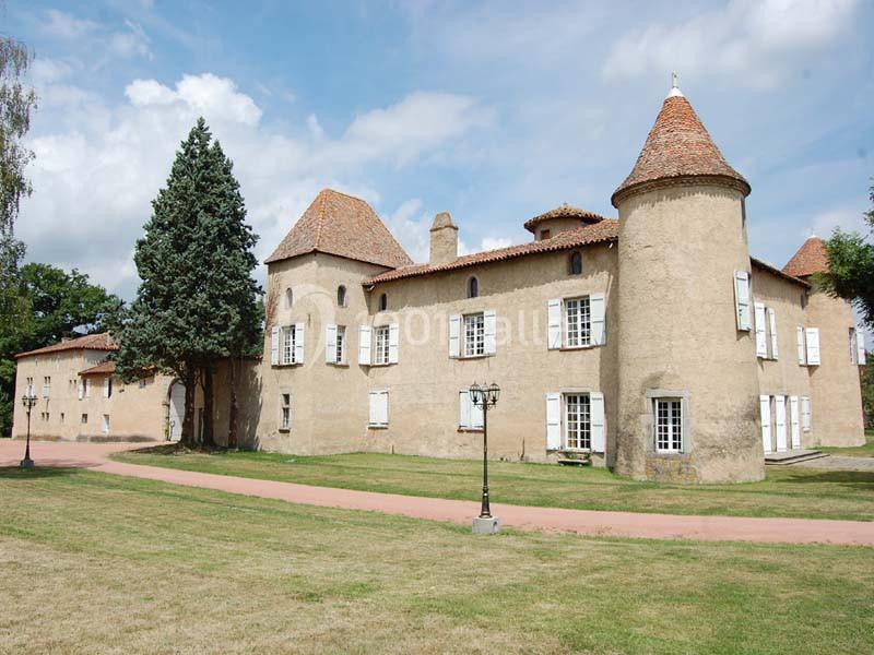 Château ancien avec tours rondes, fenêtres blanches et toit en tuiles, entouré d'une pelouse et d'un chemin.