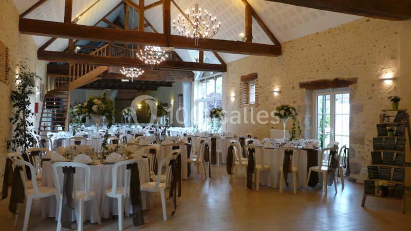 Salle de réception lumineuse avec tables rondes décorées, chaises blanches et poutres apparentes en bois.