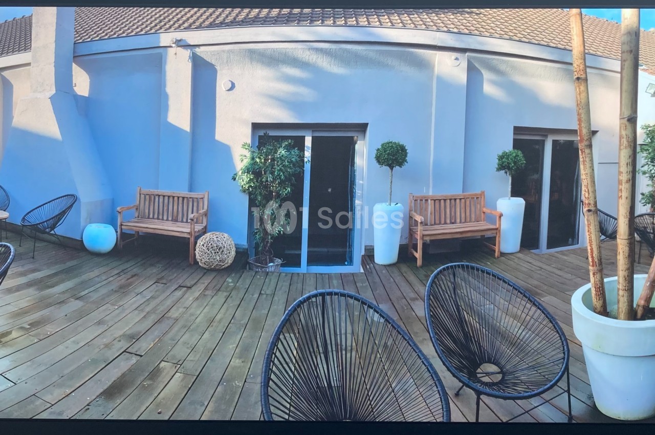 Terrasse en bois avec mobilier d'extérieur, chaises noires, bancs en bois, plantes en pots et murs blancs.