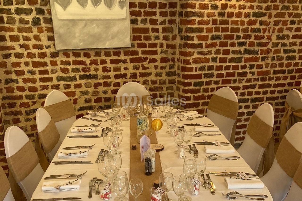 Table élégamment dressée pour un repas, entourée de chaises blanches avec nœuds beiges, devant un mur en briques.