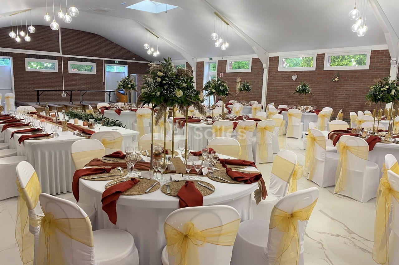Salle de réception décorée avec des tables rondes, nappes blanches, chaises ornées de rubans jaunes et centres de table…