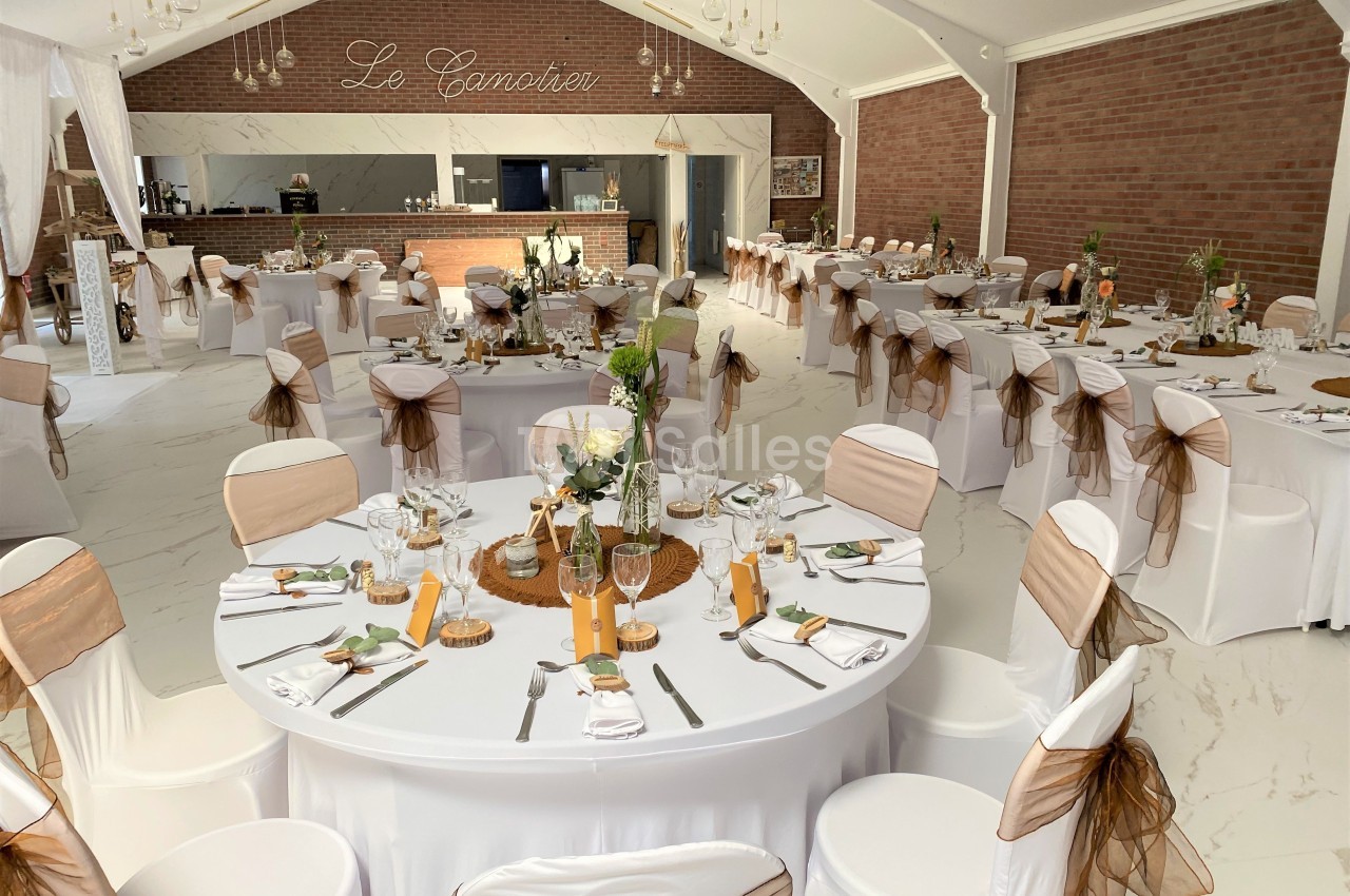 Salle de réception décorée avec des tables rondes, nappes blanches, chaises ornées de rubans et centre de table floral.