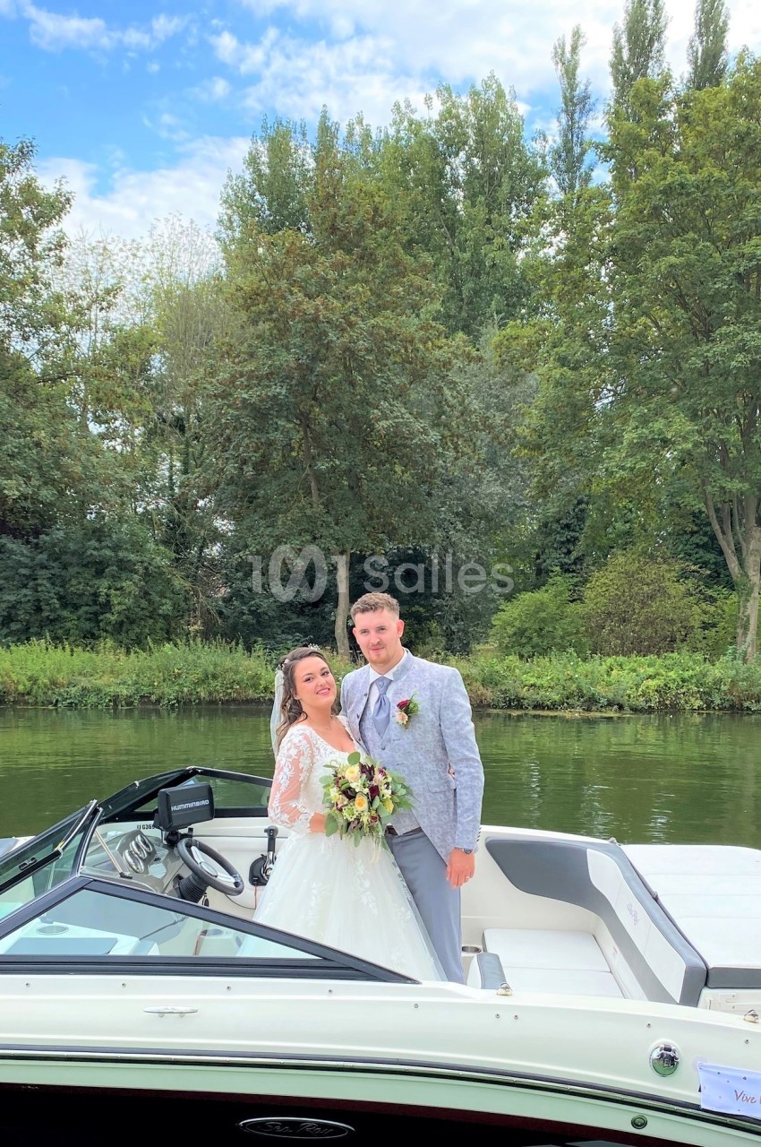 Un couple en tenue de mariage pose sur un bateau au bord d'une rivière entourée d'arbres.