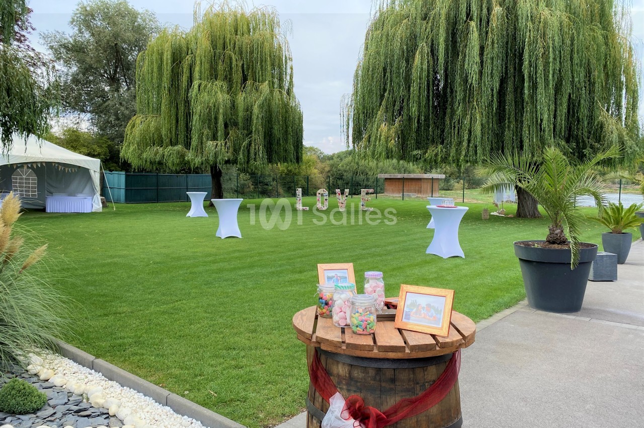 Jardin avec pelouse, tables hautes décorées, saule pleureur en arrière-plan et tonneau avec bonbonnières au premier plan.