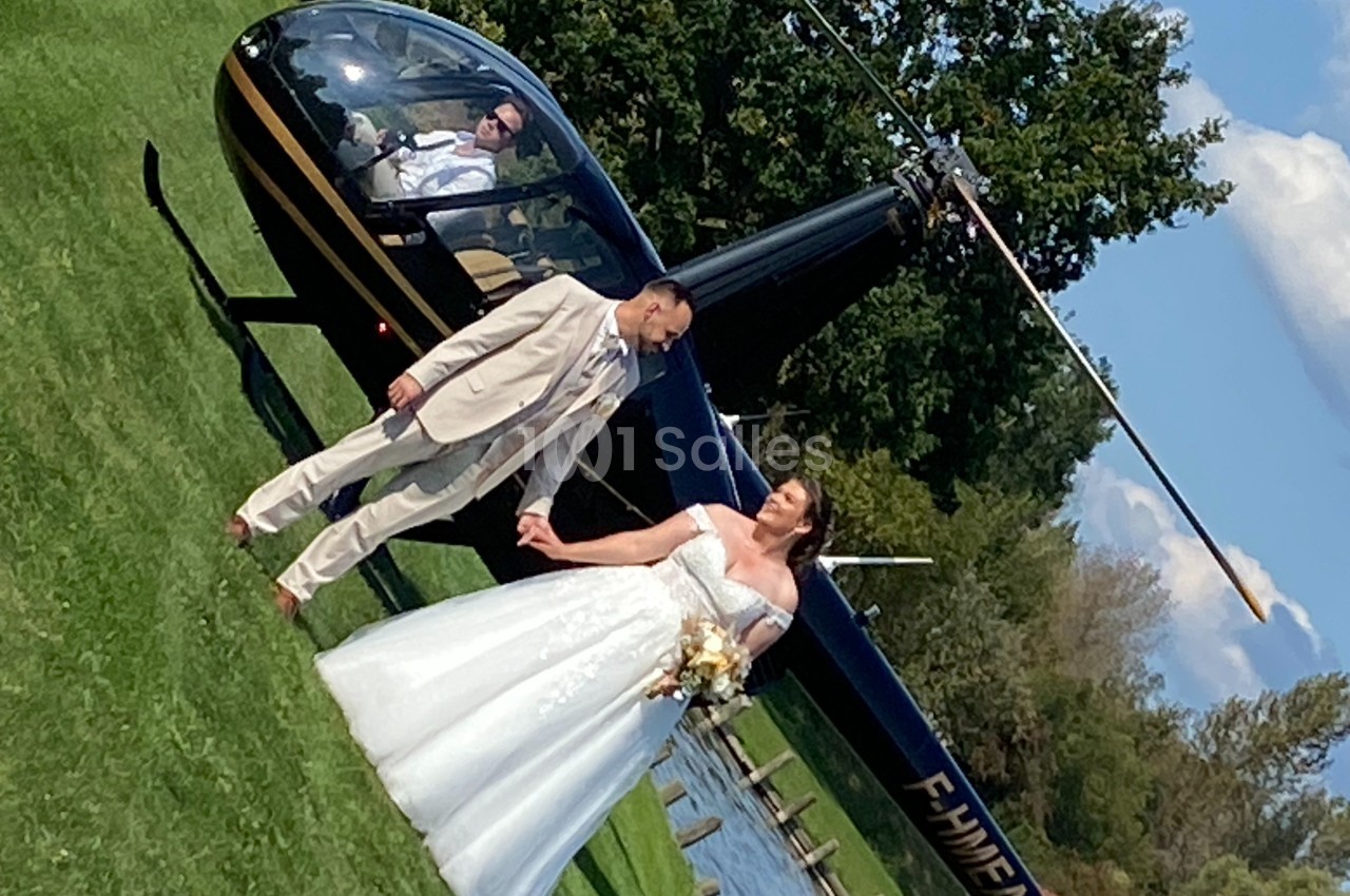 Un couple de mariés pose devant un hélicoptère noir sur une pelouse, par une journée ensoleillée.