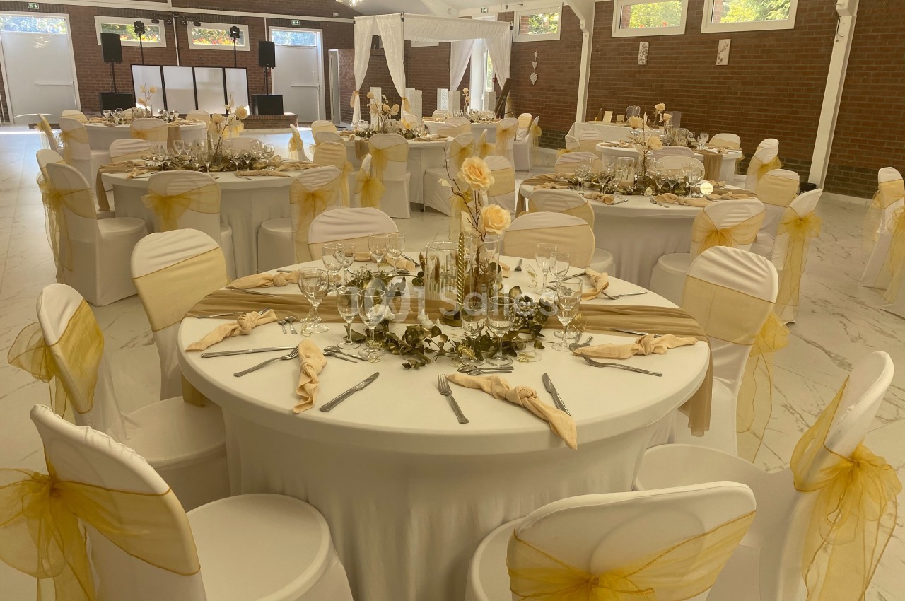 Salle de réception décorée pour un événement, avec des tables rondes ornées de nappes blanches et de chaises habillées de…