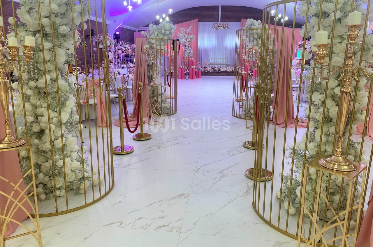 Allée décorée de fleurs blanches et de structures dorées menant à une salle de réception ornée de tons roses.