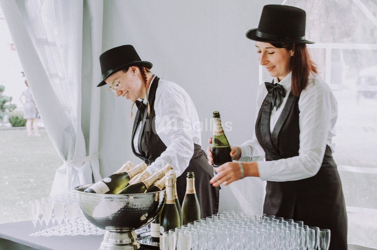 Deux serveuses en tenue formelle ouvrent des bouteilles de champagne près d'une table garnie de verres.