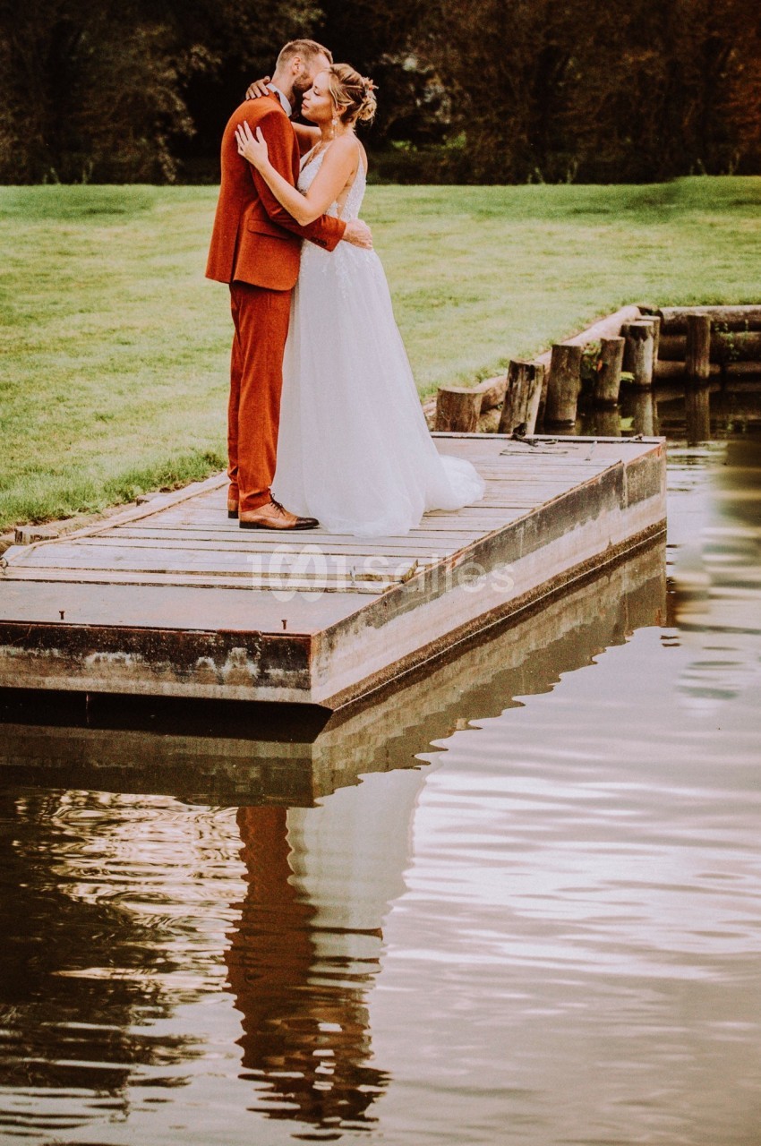 Un couple s'embrasse sur un ponton en bois au bord d'un plan d'eau, entouré de verdure.