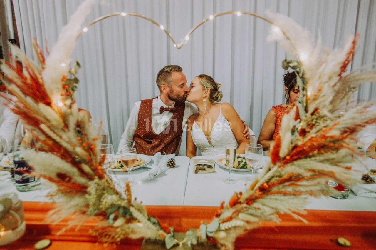 Un couple s'embrasse tendrement lors d'un repas de mariage, encadré par une décoration en forme de cœur.