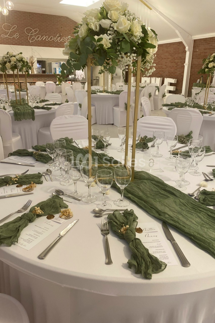 Salle de réception décorée avec des tables rondes, nappes blanches, centres de table fleuris et chemins de table verts.