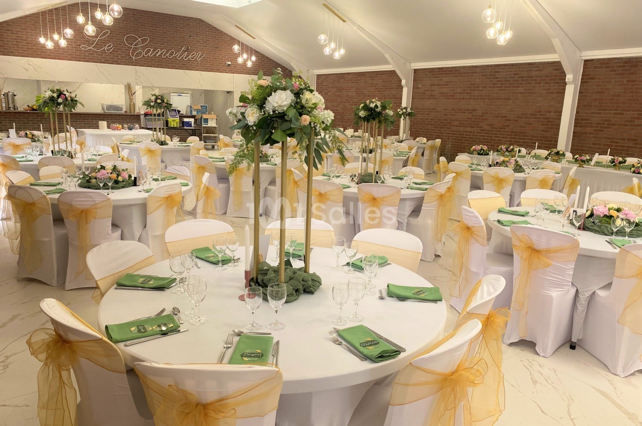 Salle de réception décorée avec des tables rondes, nappes blanches, chaises ornées de nœuds jaunes et centres de table…