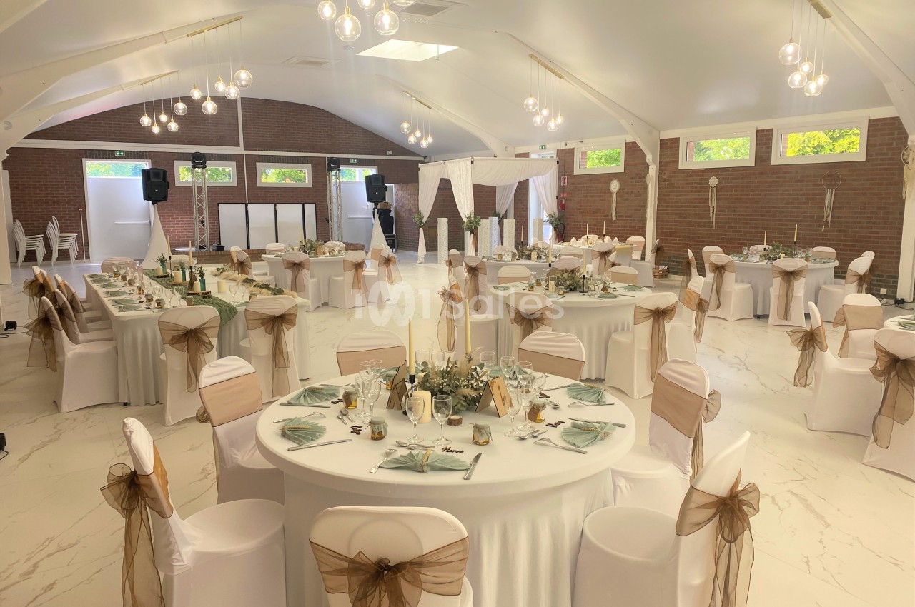 Salle de réception décorée pour un événement, avec tables rondes dressées, nappes blanches et chaises ornées de nœuds beige.