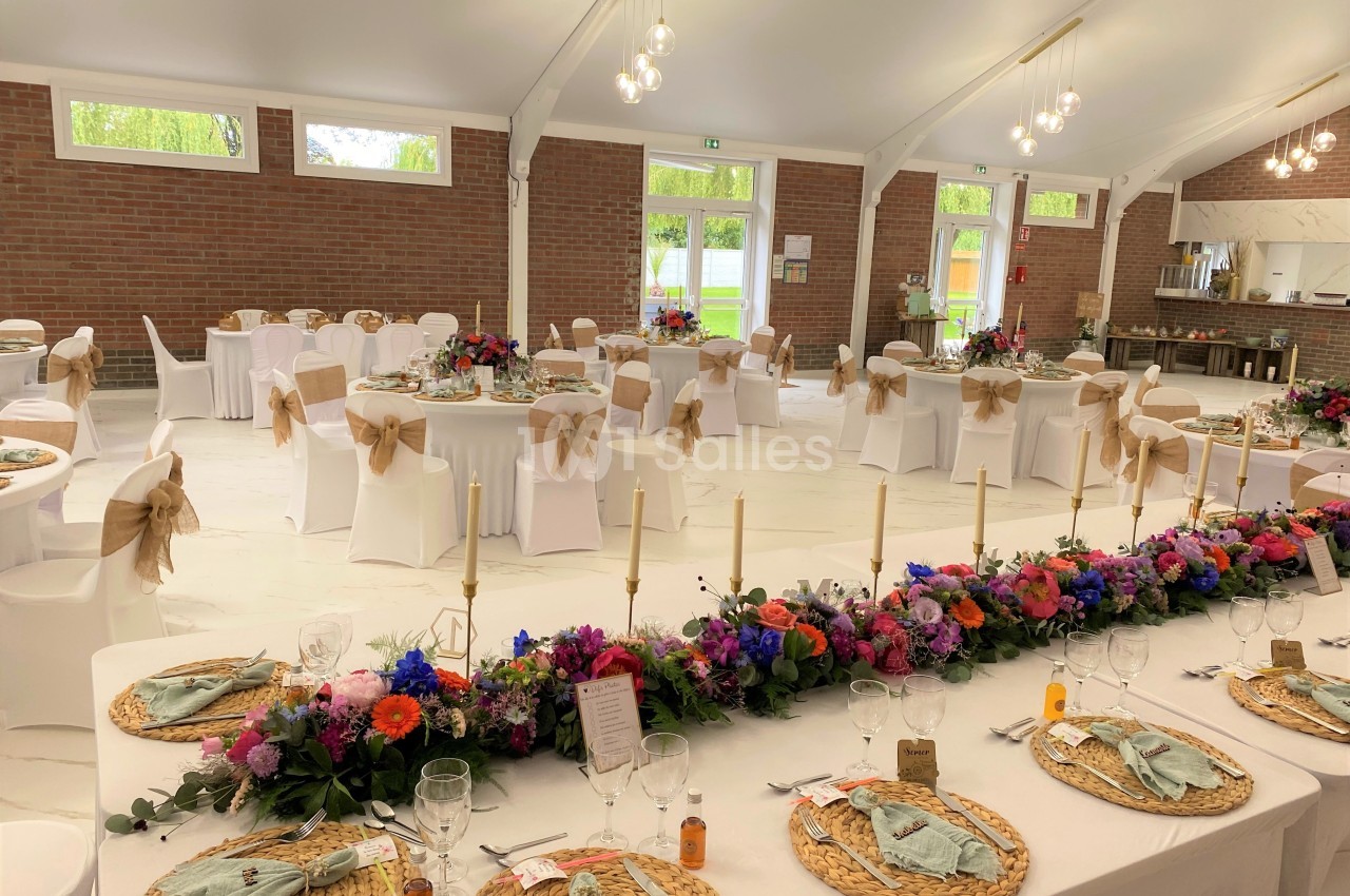 Salle de réception décorée avec des tables dressées, nappes blanches, fleurs colorées et chaises ornées de nœuds beiges.