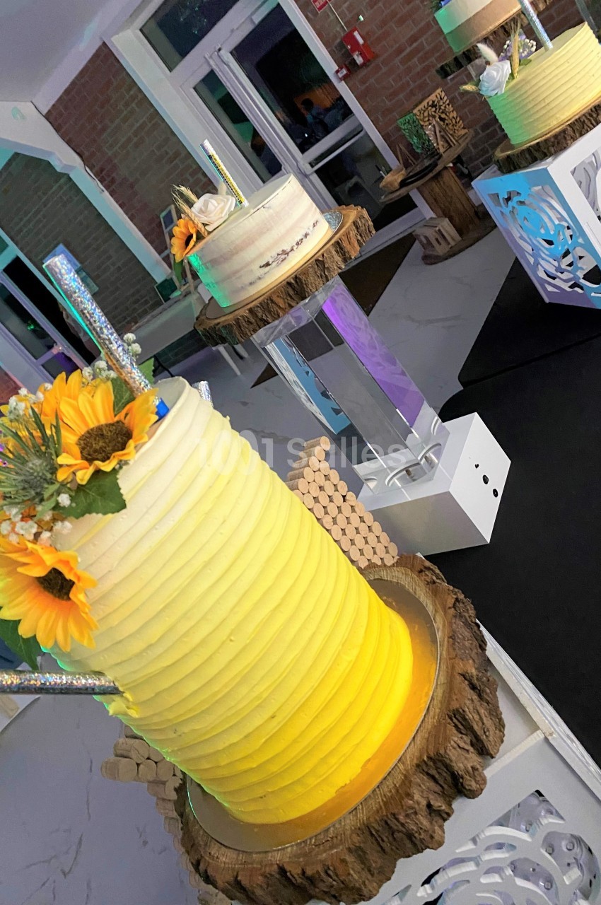 Gâteau cylindrique jaune décoré de tournesols et d'éléments brillants, posé sur une base en bois dans une salle éclairée.