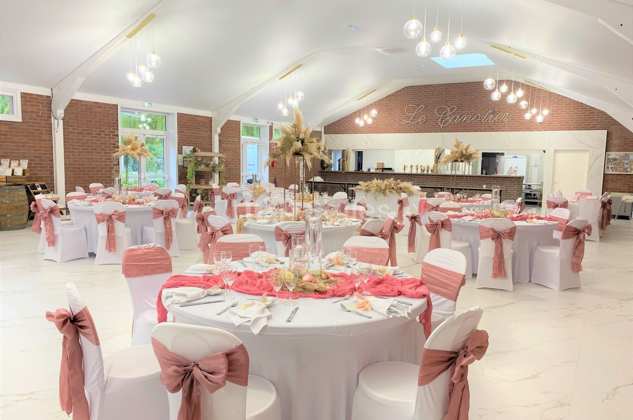 Salle de réception décorée dans des tons roses et blancs, avec tables dressées et chaises ornées de nœuds.