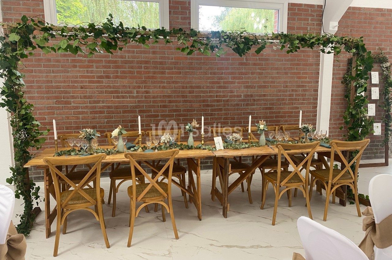 Table en bois décorée de bougies, fleurs et feuillage, entourée de chaises, sous une arche végétale dans une salle lumineuse.
