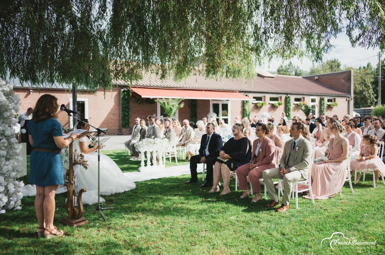 Cérémonie en plein air avec des invités assis sous un arbre, face à une mariée et une intervenante au micro.