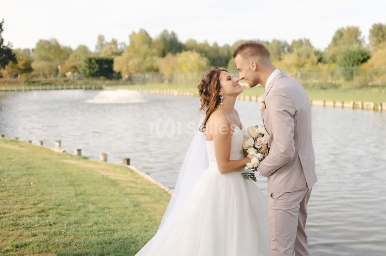 Un couple de mariés s'embrasse près d'un lac entouré de verdure par une journée ensoleillée.