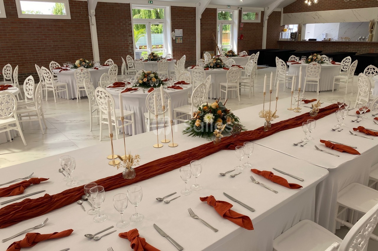 Salle de réception décorée avec des tables dressées, nappes blanches, serviettes orange et centres de table floraux.