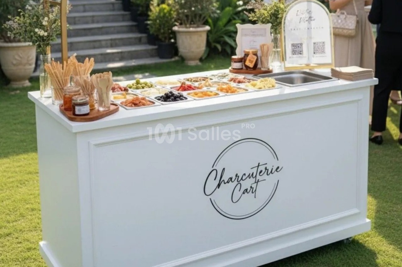 Chariot blanc avec un assortiment de charcuteries, fromages et condiments, disposé sur une pelouse extérieure.