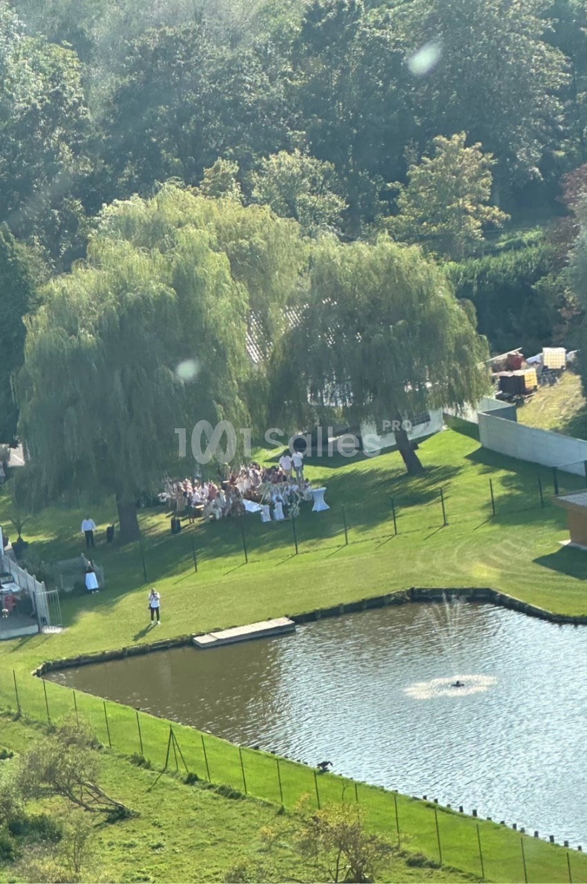 Vue aérienne d'un rassemblement en plein air sous des arbres près d'un étang avec une fontaine, dans un cadre verdoyant.