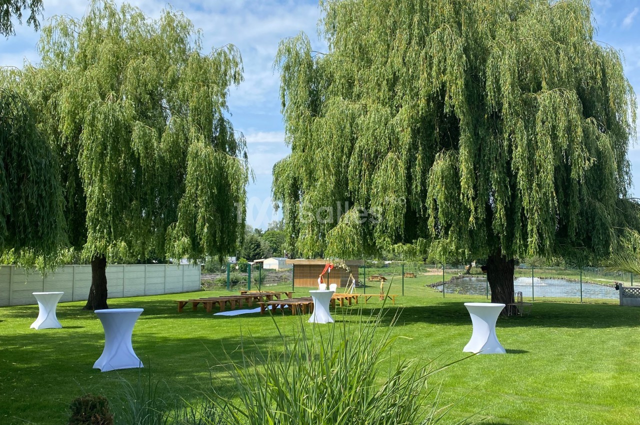 Jardin verdoyant avec deux grands saules, tables hautes blanches et bancs en bois, sous un ciel dégagé.
