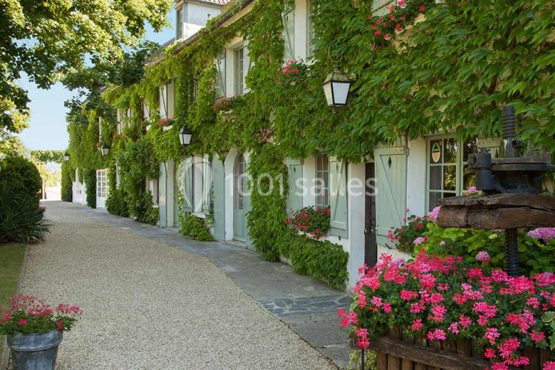 Façade de maison couverte de végétation avec volets verts, allée en gravier et fleurs colorées au premier plan.