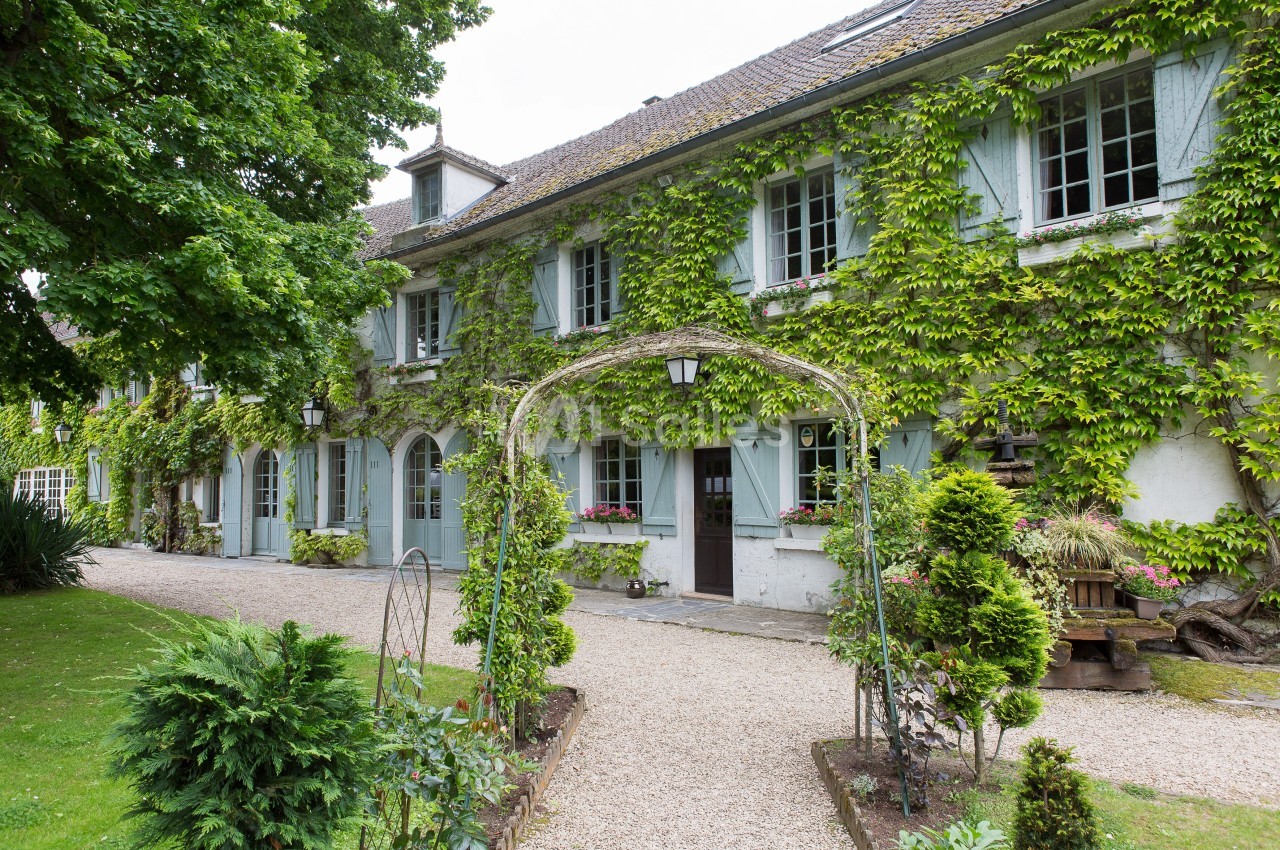Façade d'une maison ancienne couverte de lierre, avec des volets bleus et un jardin arboré à l'avant.