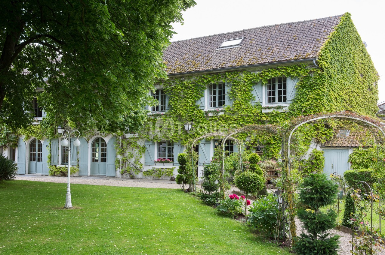Façade d'une maison traditionnelle recouverte de lierre, entourée d'un jardin verdoyant avec des arches fleuries.