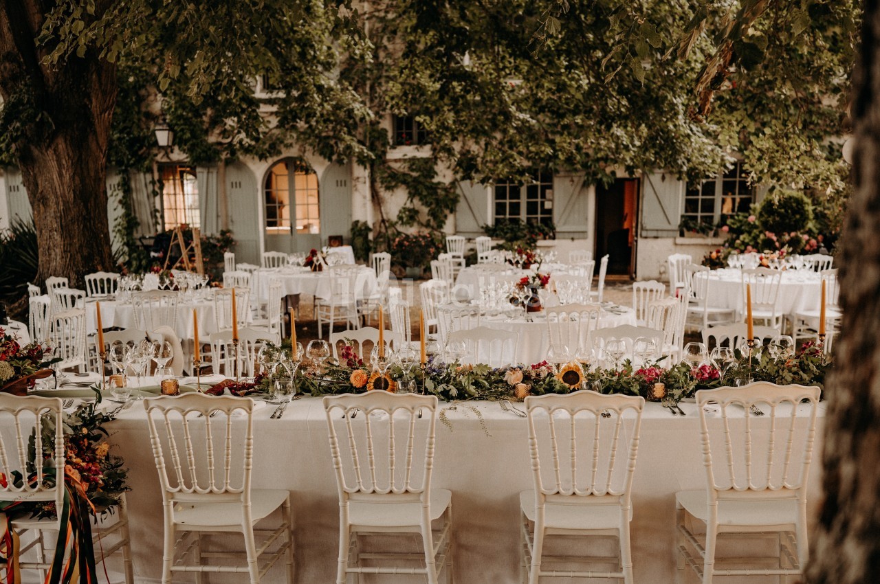 Tables de réception décorées avec des fleurs et des bougies, disposées en extérieur sous des arbres feuillus.