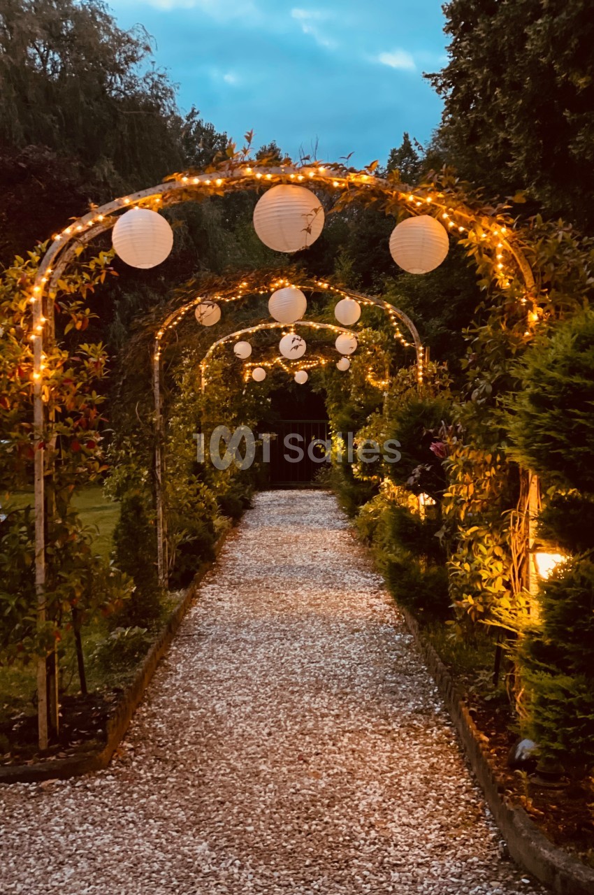 Allée de gravier bordée d'arches décorées de guirlandes lumineuses et de lanternes en papier, au crépuscule.