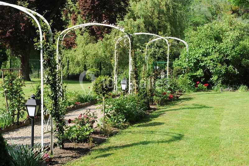 Allée de jardin bordée d'arches métalliques couvertes de plantes grimpantes, entourée de pelouse et d'arbustes.