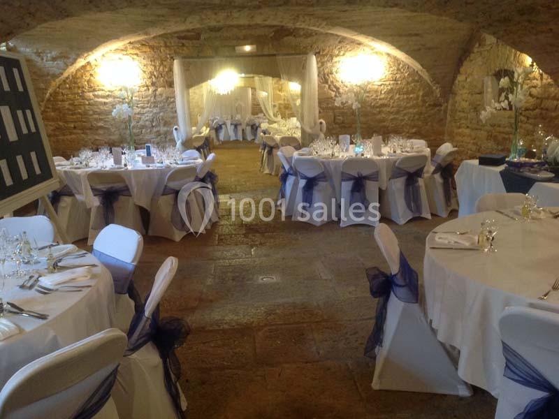 Salle voûtée en pierre aménagée pour un événement, avec tables rondes dressées et décorées de nœuds violets.