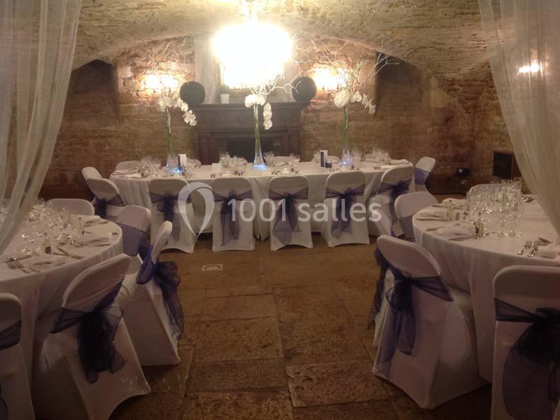 Salle voûtée en pierre aménagée pour un événement, avec tables rondes dressées et chaises décorées de rubans violets.