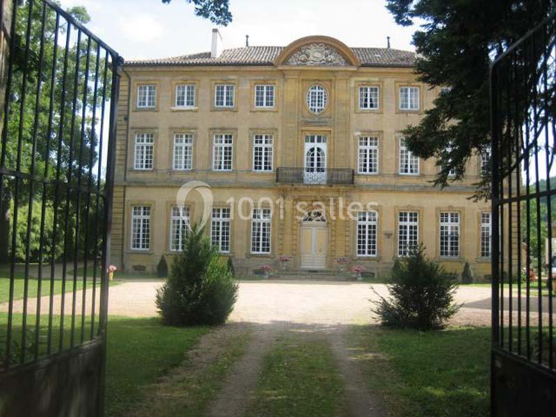Façade d'un grand bâtiment ancien en pierre jaune, entouré de verdure, vue depuis une entrée avec portail ouvert.
