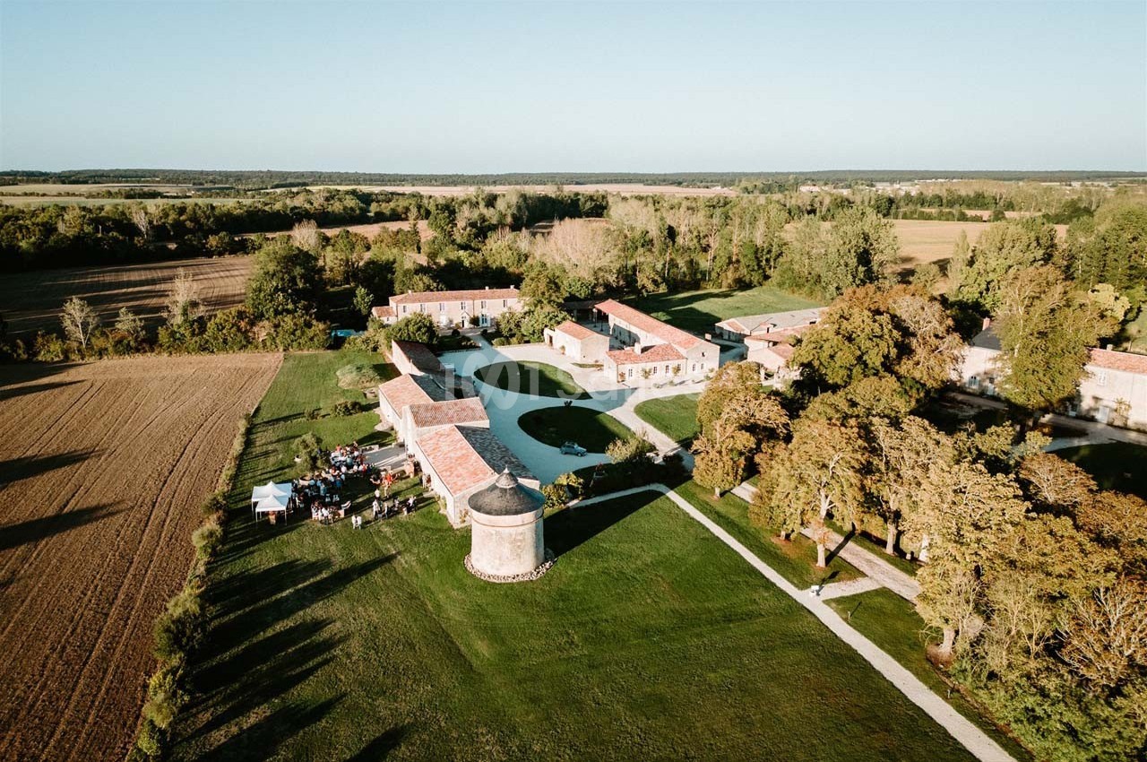 Vue aérienne d'un domaine rural avec bâtiments en pierre, pelouses, arbres et champs environnants.