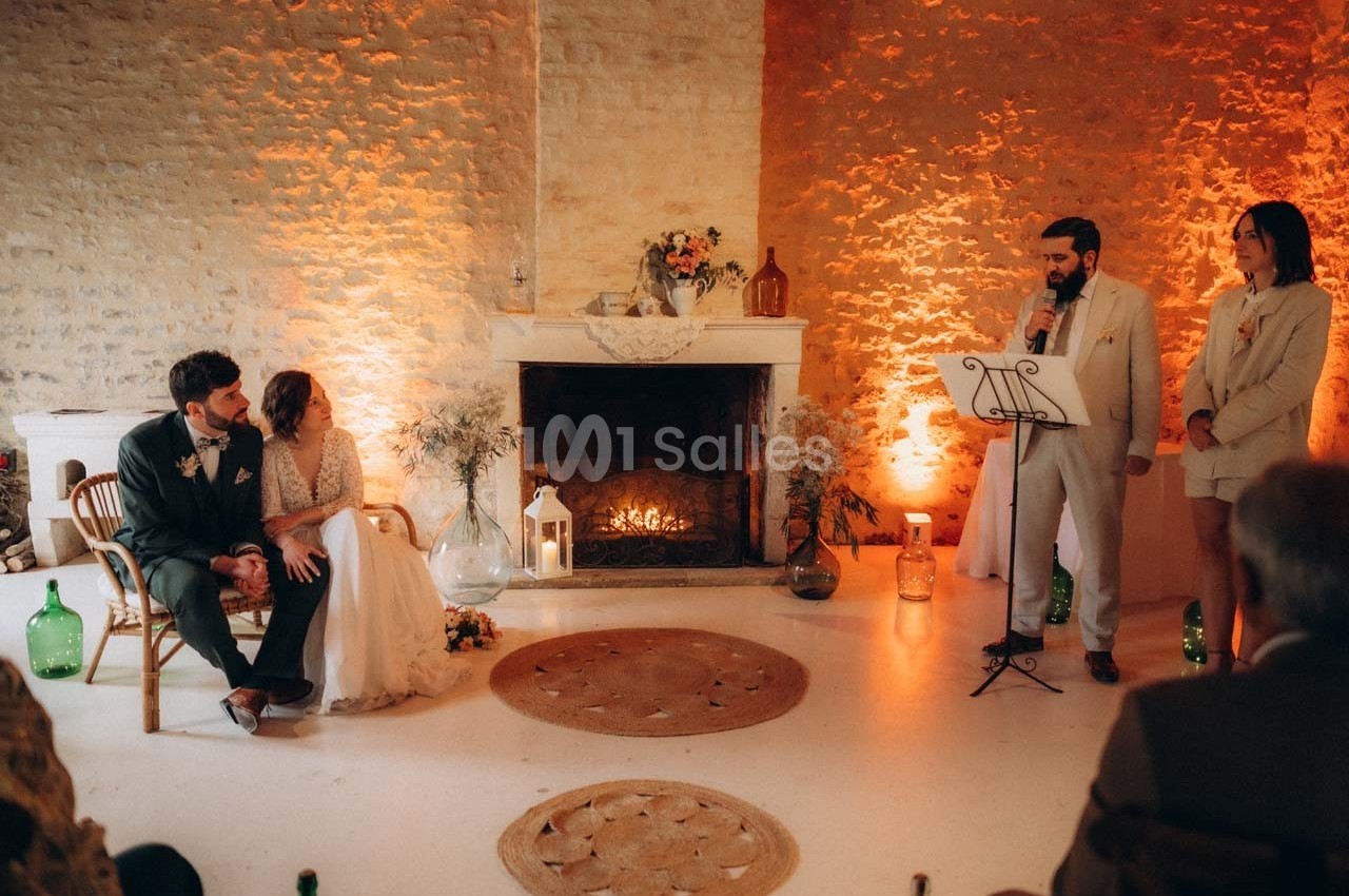 Un couple assis près d'une cheminée allumée écoute un discours lors d'une cérémonie dans une salle éclairée chaleureusement.