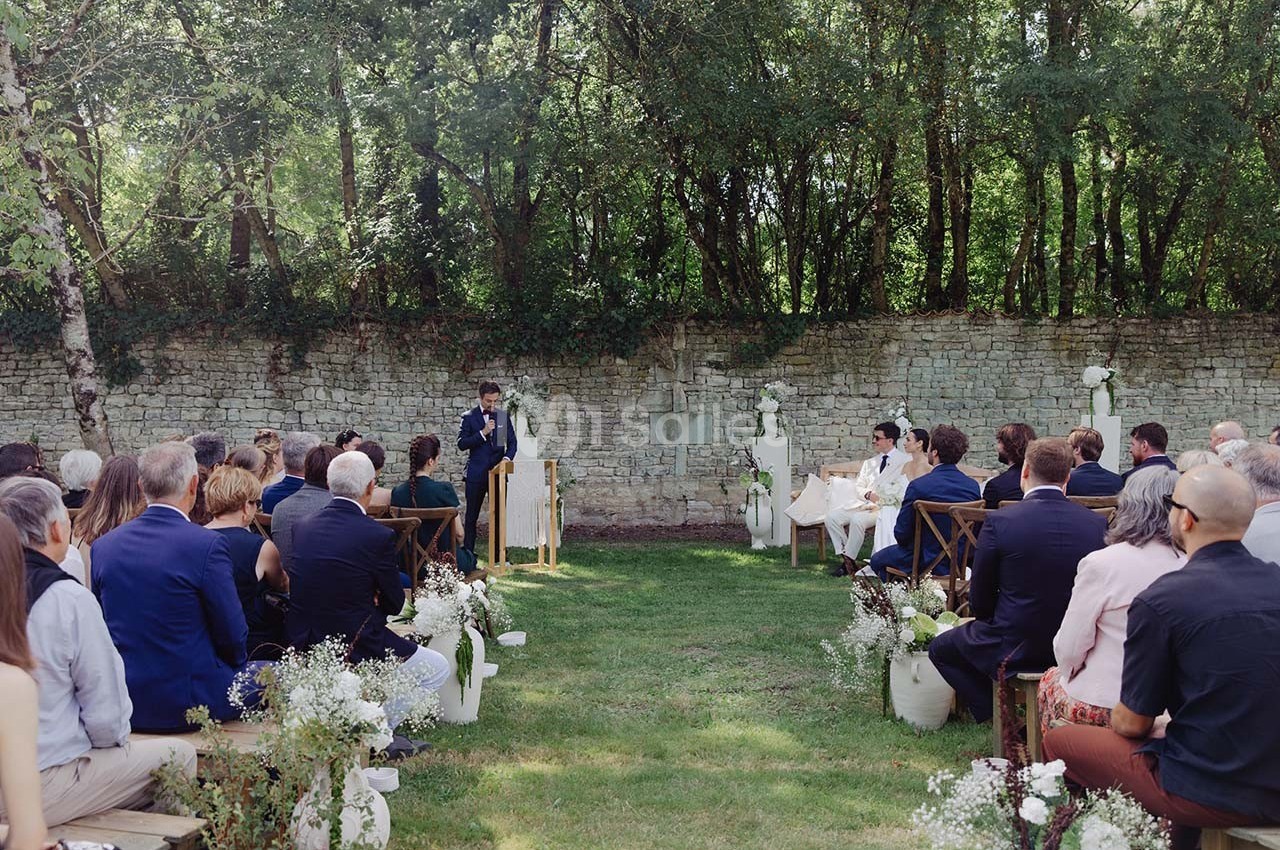 Cérémonie de mariage en plein air avec des invités assis, un officiant et les mariés devant un mur en pierre.