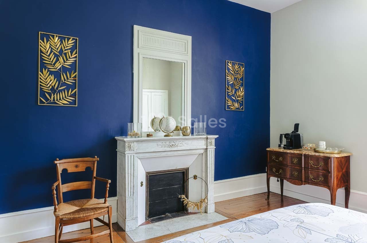 Cheminée blanche avec miroir, mur bleu décoré de cadres dorés, meuble en bois et chaise dans une chambre lumineuse.