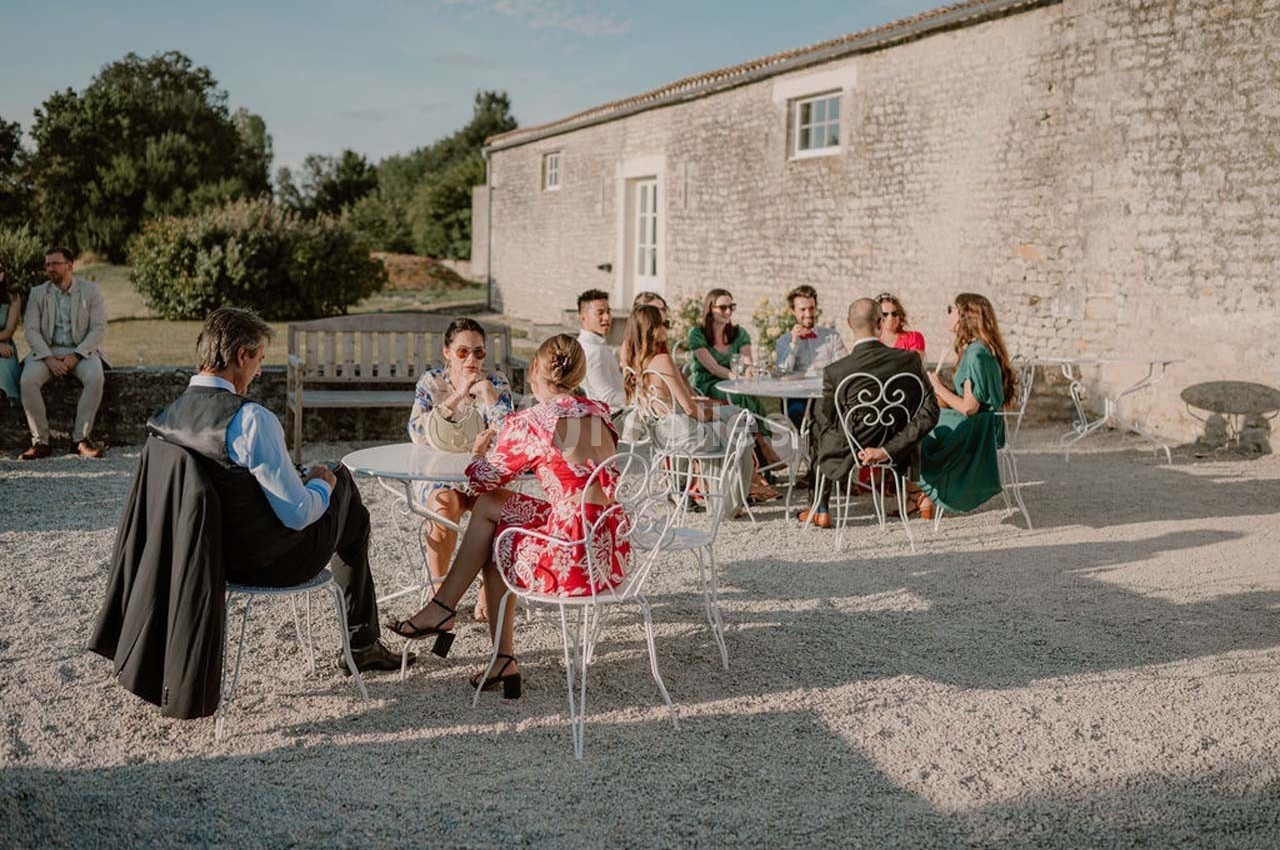 Personnes assises à des tables en plein air devant un bâtiment en pierre, discutant par une journée ensoleillée.